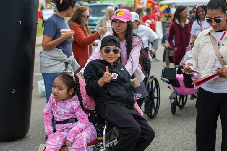 a kid in sunglasses give a thumbs up while sitting on the arm of a wheelchair with a young girl in it