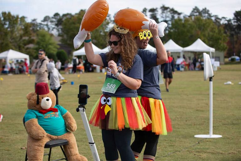 two women in turkey skirts holding inflatable turkey legs pose for a picture with a stuffed Fezzy